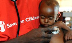 A faceless man in a red Save the Children t-shirt holds a stethoscope to a child's heart as he holds the child in his arms.