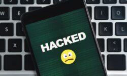 A cellphone screen says HACKED with a frown emoji under the word HACKED. The background is a keyboard.
