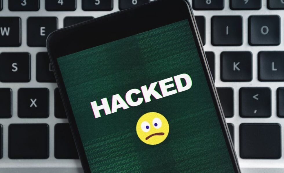 A cellphone screen says HACKED with a frown emoji under the word HACKED. The background is a keyboard.